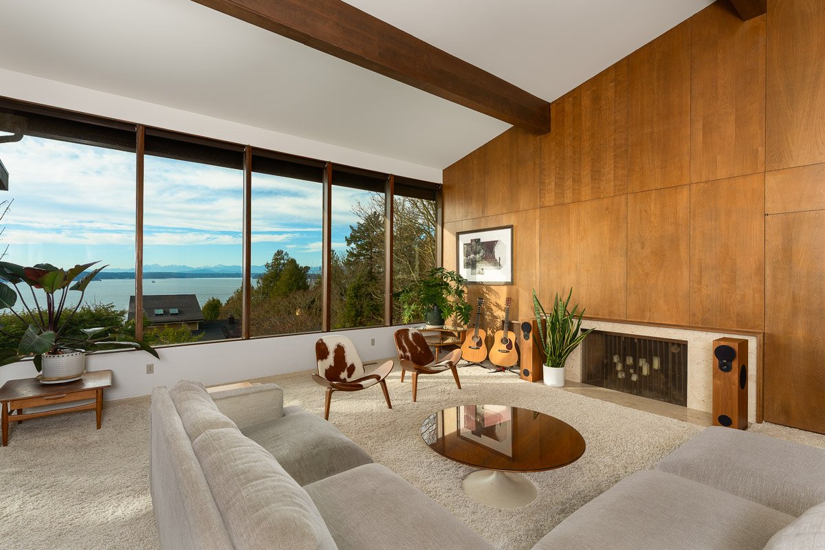 Professional real estate photography of a mid-century Seattle living room with wood-paneled walls, expansive windows, a fireplace, and stunning water views.