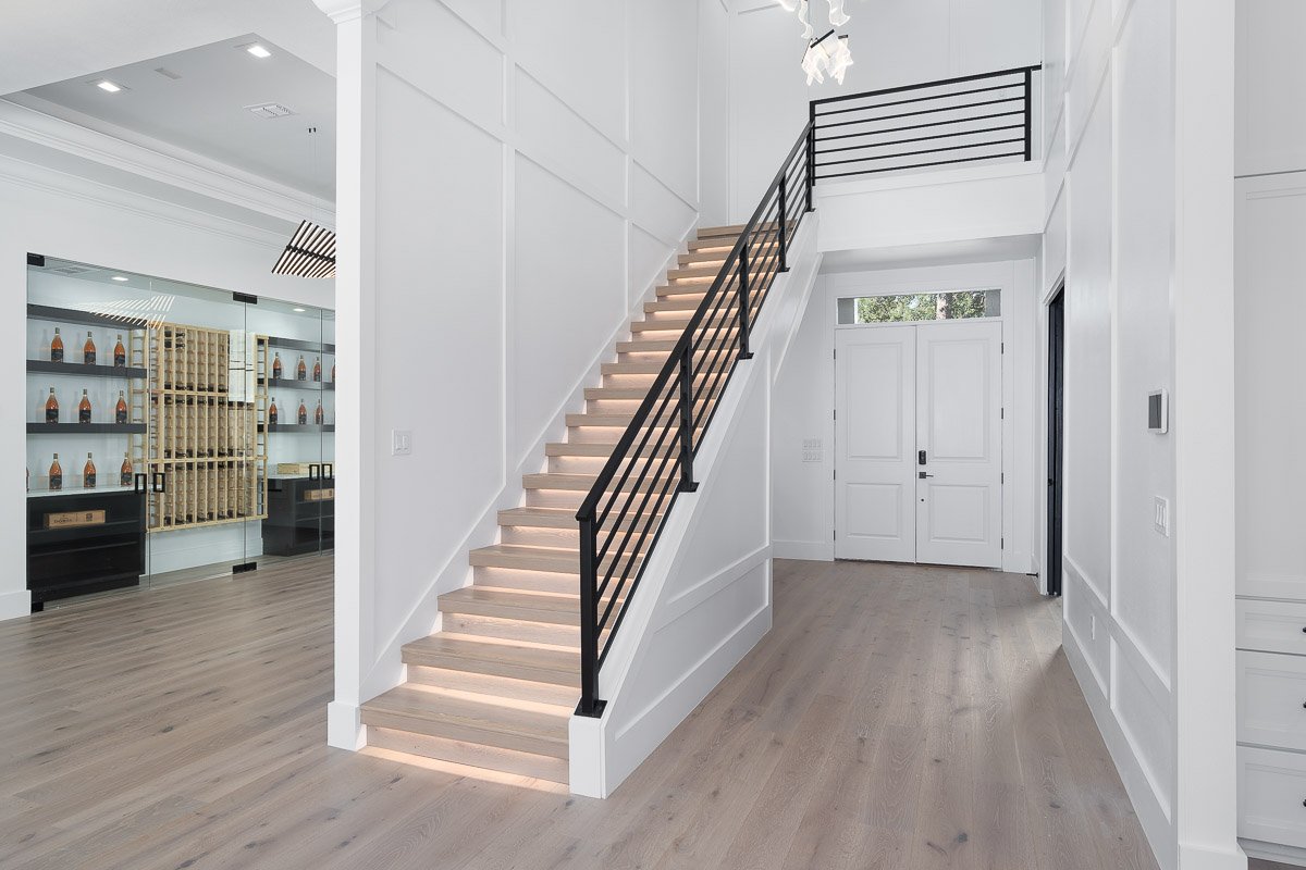 Professional real estate photography of a luxury Seattle entryway with a modern staircase, illuminated steps, black railings, and custom wine storage.
